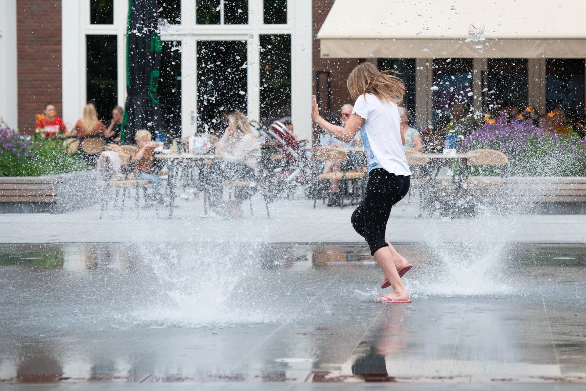 Spektakelfontein Kerkplein Hoogeveen 2 Een meisje rent door het spiegelende water op het Kerkplein in Hoogeveen, met een terras op de achtergrond