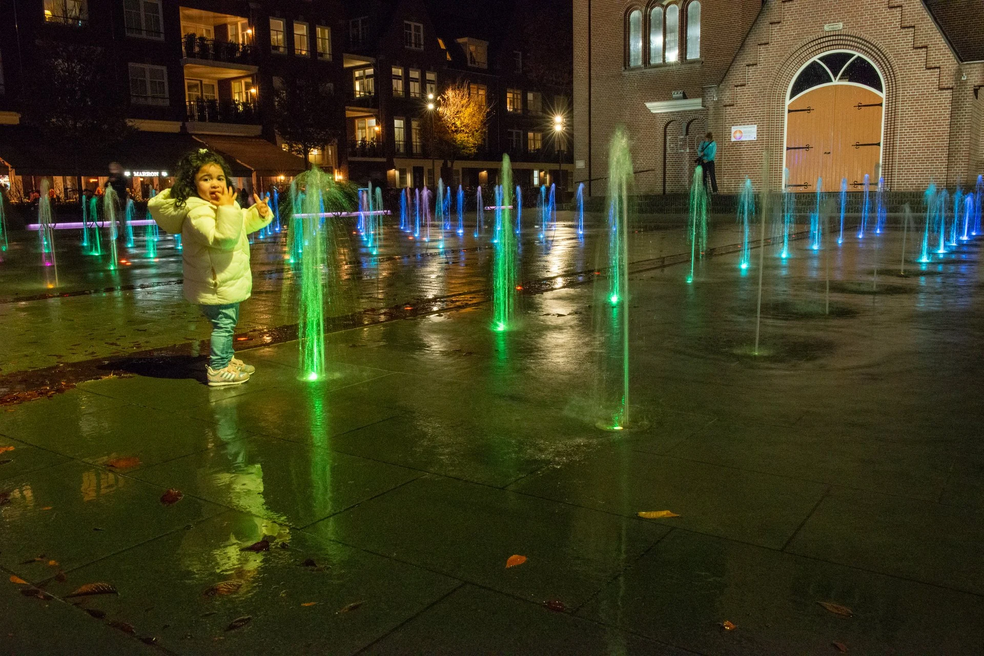 Een kind staat tussen groen en blauw verlichte fonteinen op het Kerkplein in Hoogeveen in de avond