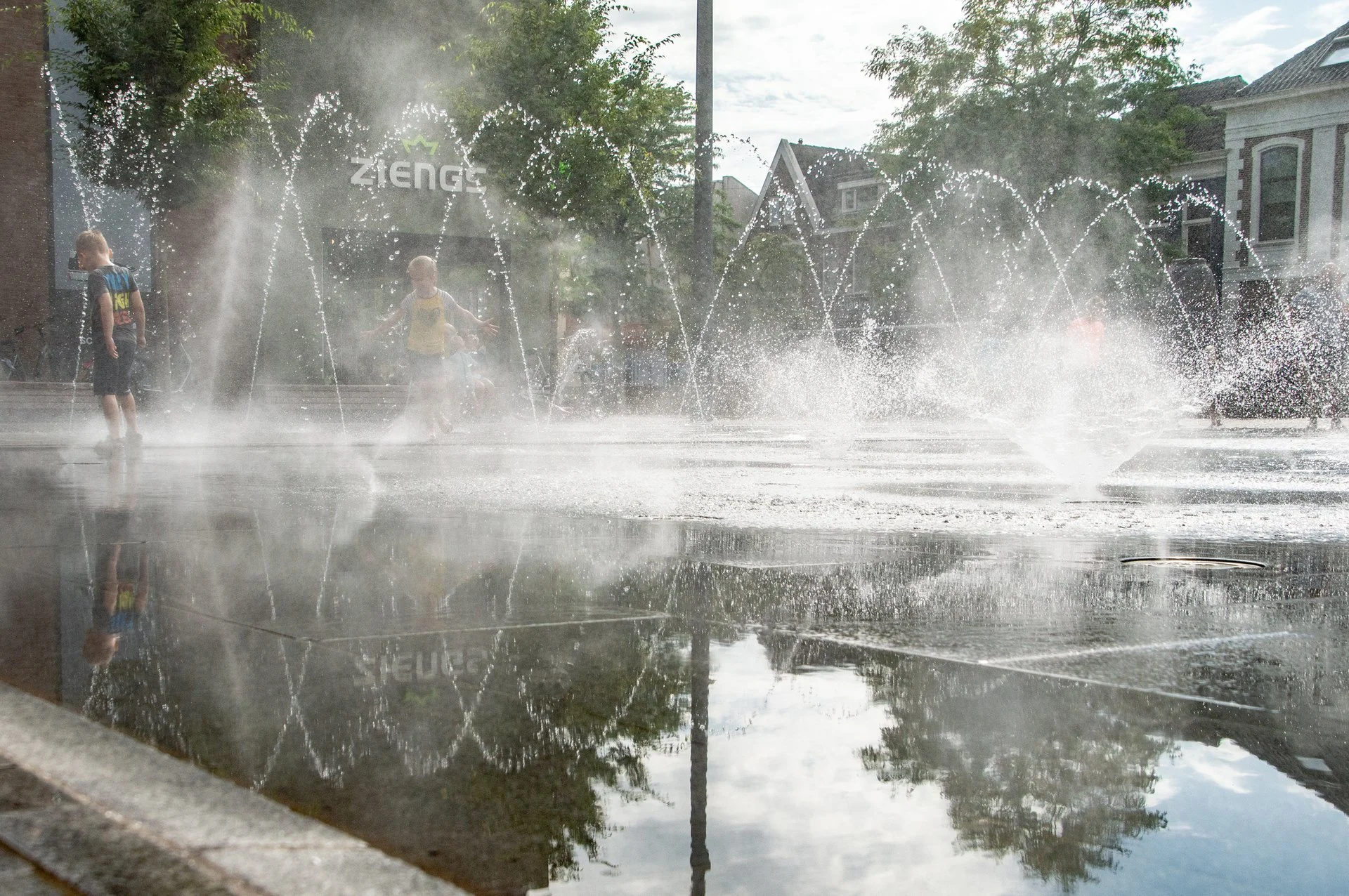 Fonteinen en nevel boven het spiegelende water op het Kerkplein in Hoogeveen, met bomen en gebouwen erachter