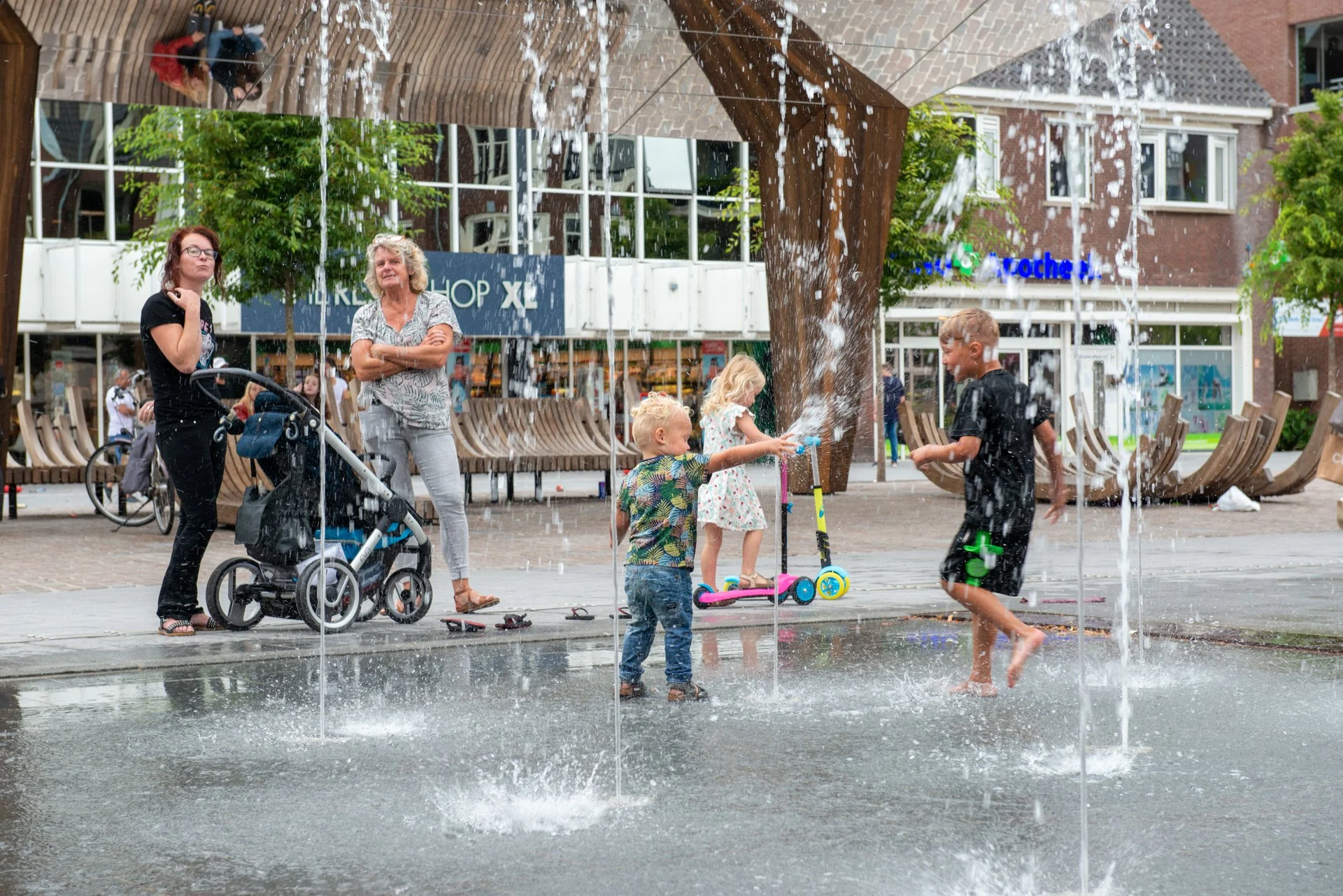 Ouders en kinderen bij de fonteinen op het Kerkplein in Hoogeveen, met de spiegelkraan op de achtergrond