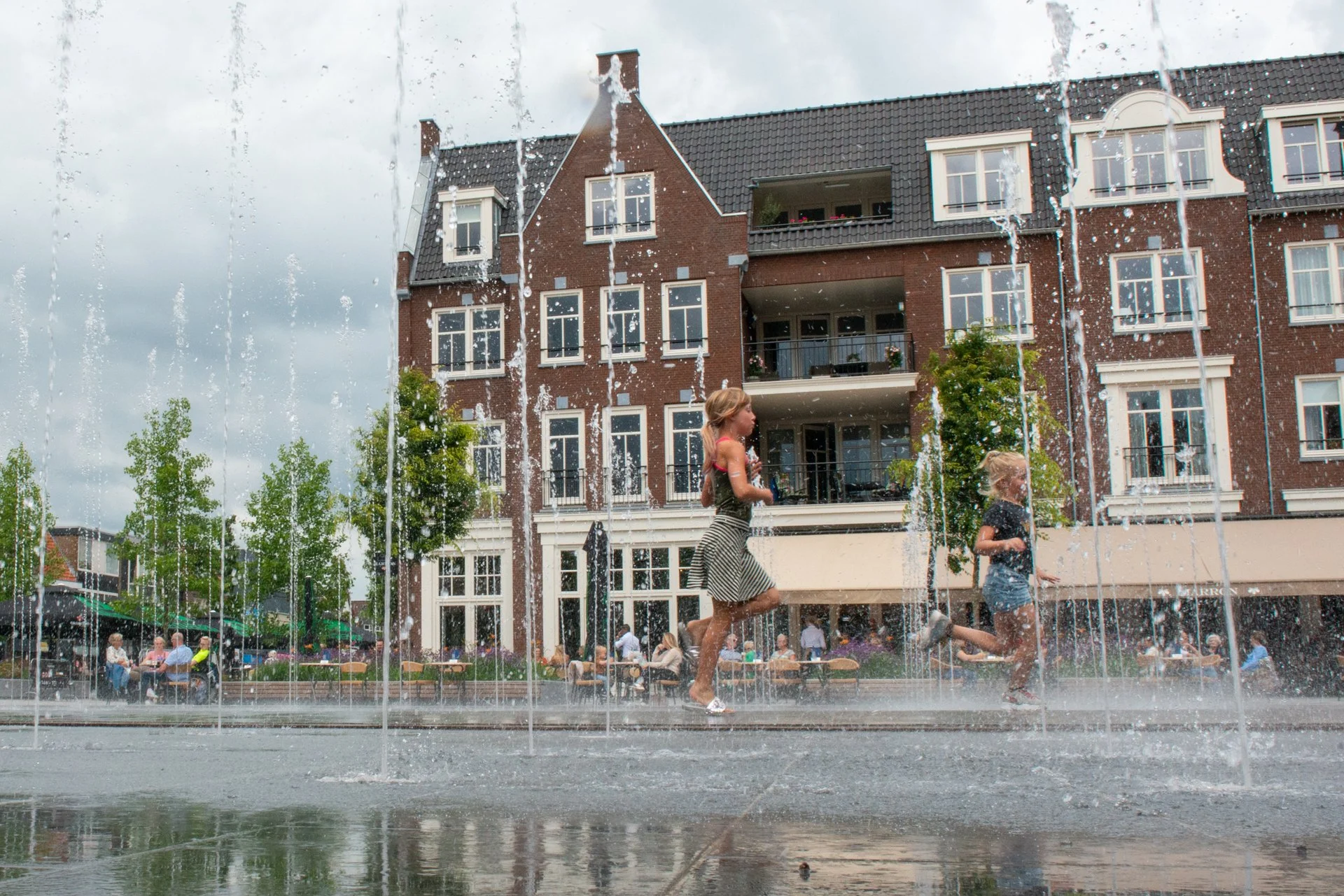 Kinderen rennen langs de fonteinen voor de gevels met terras op het Kerkplein in Hoogeveen